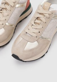 Sneakers beige realizzati in suede e mesh, con dettagli dorati, una suola nera e un tallone verde e arancione. Punta arrotondata e design con lacci.