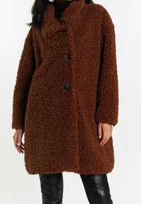 Brun, shaggy tekstureret frakke med frontknaplukning, stor krave og løs pasform. Sorte knapper kontrasterer med det bløde materiale.