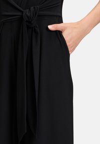 Zwarte jumpsuit met een detail van een knoop aan de voorkant, zachte stof en zijzakken, die een schone en gestroomlijnde uitstraling benadrukt.