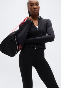 Μαύρο αθλητικό μπουφάν με φερμουάρ και μακριά μανίκια, σετ με εφαρμοστά κολάν, συνδυασμένο με μαύρη τσάντα duffel με κόκκινες και λευκές ρίγες.