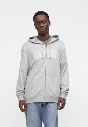 Joven con sudadera con capucha gris claro y cremallera con el texto "GAP 1969" y vaqueros azules, de pie sobre un fondo blanco liso.