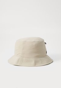 Beige bucket hat with a round crown and wide brim. Features vertical stitching and a small adjustable cord with a toggle. Smooth texture.