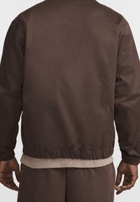 Brown lightweight jacket with a relaxed fit, elasticated cuffs, and waistband. Smooth texture with minimal stitching detail.
