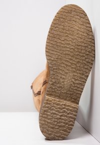 Sula av en beige mockaboot med texturerad gummisula och blixtlås på sidan, uppställd mot vita hörnväggar.