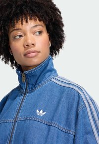 Veste en denim avec col montant, fermeture zippée et accents blancs à trois bandes sur les épaules. Logo notable sur la poitrine.