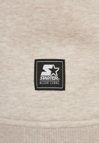Tela beige con una textura suave que presenta un parche de logo cuadrado negro. El parche muestra "STARTER" y "BLACK LABEL" en letras blancas en negrita.