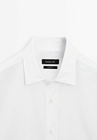 Chemise blanche à col pointu, boutons supérieurs fermés et étiquette noire Massimo Dutti visible à l'intérieur du col.