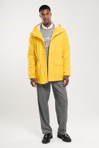 Veste de pluie jaune imperméable avec capuche, fermeture à bouton-pression et deux poches avant, superposée à un pull en gris et un pantalon gris.