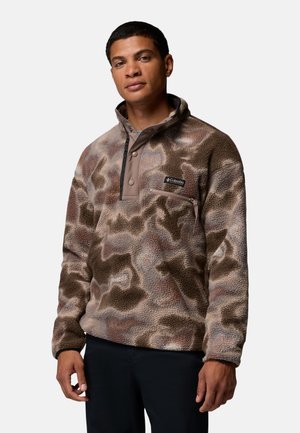 Man wearing a brown and gray camouflage patterned fleece pullover with a buttoned collar and chest pocket, standing against a plain background.