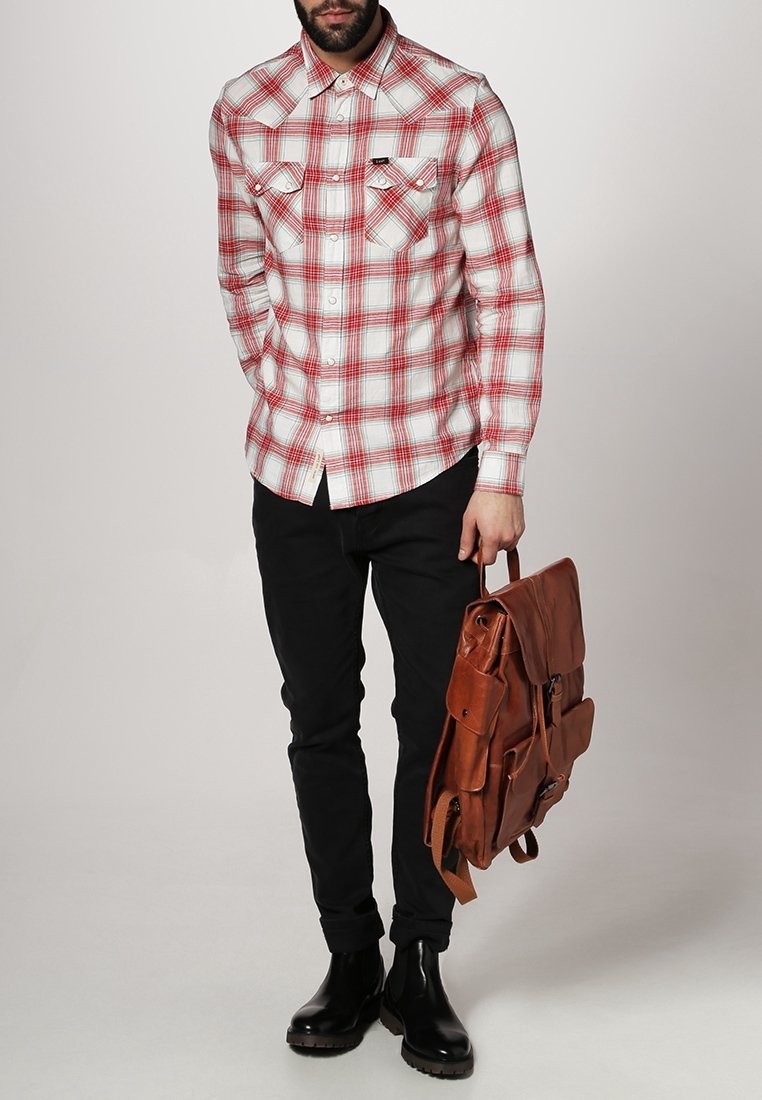 Homme portant une chemise à carreaux rouge et blanche, pantalon noir, bottes noires, tenant un sac à dos en cuir marron par sa poignée supérieure.