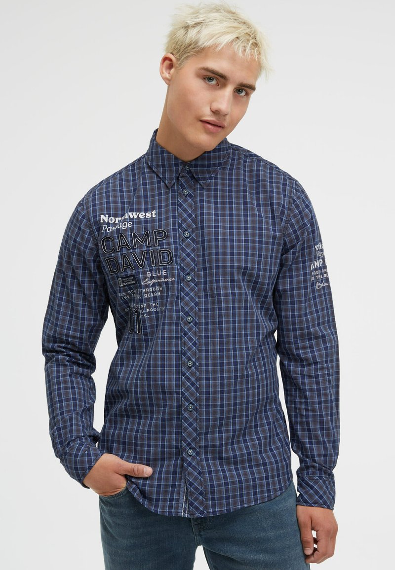 Blau kariertes langärmliges Hemd mit Button-Down-Kragen, ausgestattet mit gesticktem Text und einer Taschenriegelung auf der linken Seite.