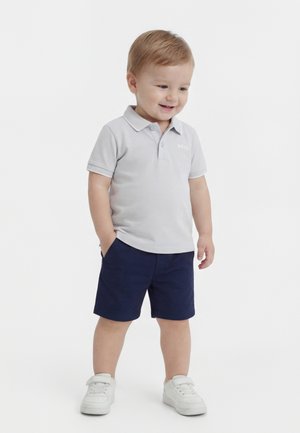 Bambino sorridente, indossa una polo grigio chiaro, pantaloncini blu navy, scarpe da ginnastica bianche, in piedi con le mani nelle tasche davanti a uno sfondo bianco semplice.