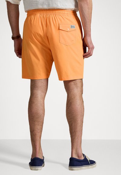 Pantaloni scurți portocalii cu talie elastică, buzunar posterior cu închidere cu nasture și o textură netedă. Încălțămintea este din pânză albastră închisă.