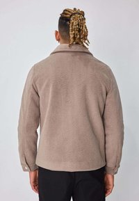 Veste en polaire beige à finition texturée, dotée d'un col pointu et de poignets boutonnés, vue de dos sur un fond neutre.