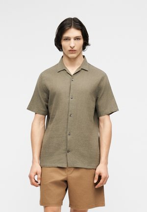 Νέος άνδρας που στέκεται με το βλέμμα προς τα εμπρός, φοράει κοντομάνικο πουκάμισο χακί με κουμπιά και ανοιχτόχρωμο καφέ σορτς μπροστά σε απλό φόντο.