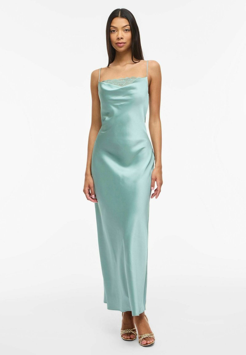 Femme portant une robe nuisette en satin bleu sarcelle avec de fines bretelles et un détail en dentelle au niveau du décolleté, debout devant un fond blanc uni.