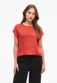 Rød sateng bluse med korte ermer, rund hals og rullede mansjetter, kombinert med svarte bukser. Glatt tekstur og lett utseende.