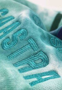 Textil bordado con letras turquesa sobre un fondo suave y texturizado. Presenta un diseño en degradado con tonos variados de azul.