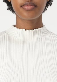 White ribbed top with a high, frayed neckline. The fabric displays vertical lines and a soft texture, close-fitting design, and minimal detailing.
