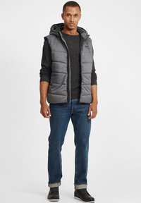 Grau gesteppte Weste mit Kapuze über einem dunklen Pullover. Dazu blaue Jeans und schwarze Schuhe. Flache Stofftextur und gestepptes Muster.