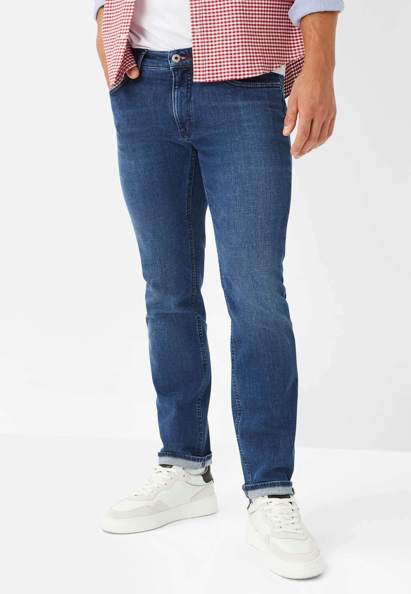 Männer Jeans aus blauem Denim, gerade geschnitten, mittlere Waschung, mit Vorderaschen und Knopfverschluss, kombiniert mit weißen Sneakern.