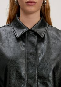 Zwarte faux leren jas met een puntige kraag, zichtbare textuur en een voorrits. Heeft schone lijnen en een gestructureerde silhouet.