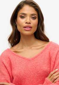 Korale sweter oversize z szerokim dekoltem w kształcie litery V i prążkową teksturą. Modelka ma długie falowane włosy i nosi małe kolczyki w kształcie obrączek.