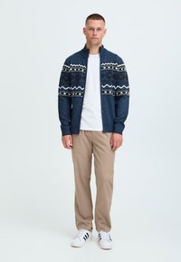 Jūrinės mėlynos megztos kardiganas su Fair Isle raštu balta ir juoda spalvomis, užtraukiamas, dėvimas ant baltos marškinėlių ir derinamas su smėlio spalvos kelnėmis bei baltais sportbačiais.