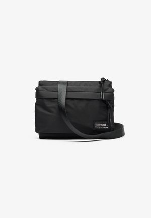 Bolsa preta compacta com uma alça ajustável, apresentando um fecho com zíper e tecido texturizado. Um rótulo indica detalhes de design e inclusão.