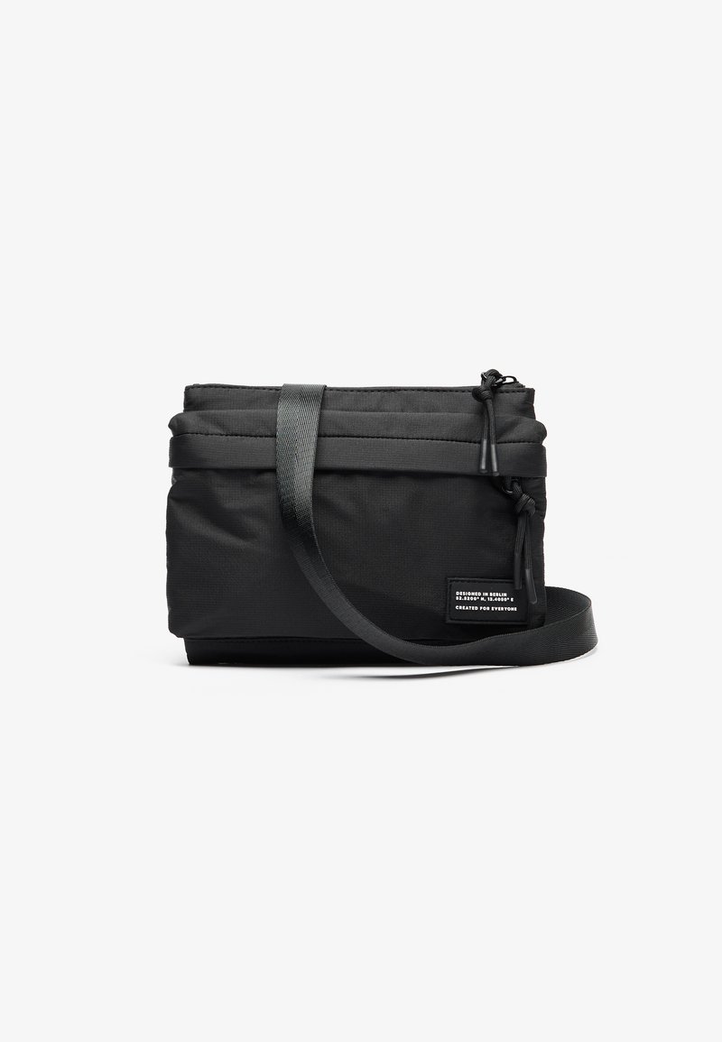 Borsa nera compatta con tracolla regolabile, dotata di chiusura con zip e tessuto testurizzato. Un'etichetta indica i dettagli del design e l'inclusività.