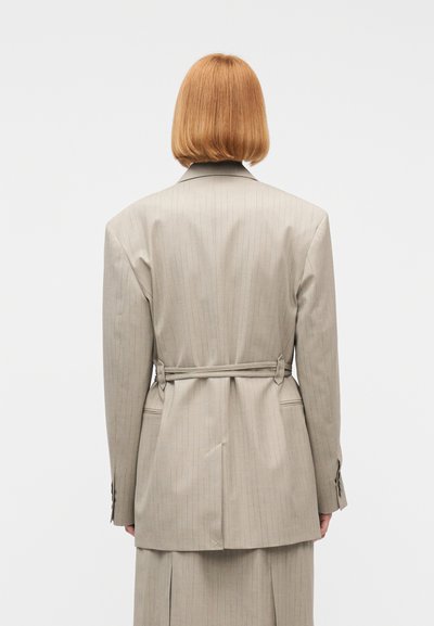 Άτομο με κοντά κόκκινα μαλλιά, ντυμένο με μπεζ σακάκι και φούστα με ρίγες, στέκεται με την πλάτη προς το μέρος μας μπροστά σε ένα απλό φόντο.