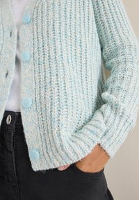 Ljusblå stickad cardigan med ribbat mönster, som har två ljusblå knappar och är kombinerad med mörka byxor.