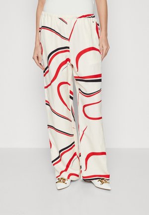 Mujer con pantalones blancos sueltos con rayas curvas abstractas en rojo y negro, combinados con zapatos blancos puntiagudos con cadenas doradas.