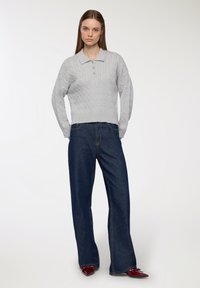 Kobieta ubrana w jasnoszary sweter w sploty z kołnierzykiem, ciemne dżinsy z szerokimi nogawkami oraz błyszczące bordowe czółenka, stojąca na tle białym.