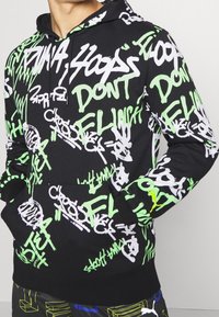 Felpa nera con cappuccio, caratterizzata da testo in stile graffiti bianco e verde neon, con cordoncino e tasca a marsupio frontale.