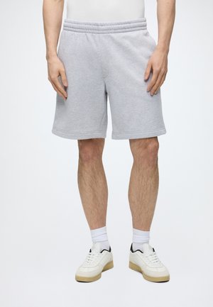 Mann trägt hellgraue knielange Sweatshorts, weiße Sneakers mit beigen Sohlen und weiße Socken vor einfarbigem Hintergrund.