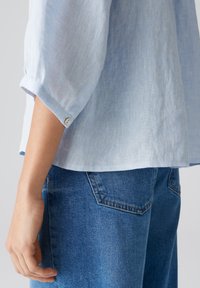 Lichtblauwe linnen overhemd met mouwen tot de elleboog en een knoopdetail bij de manchette, gecombineerd met blauwe denim jeans met een achterzak.