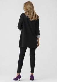 Musta tunika pidemmällä takapäällä, yhdistetty mustiin legginsseihin. Varsissa on harmaat raidalliset hihansuut; violetit korkokengät tuovat väriä asuun.