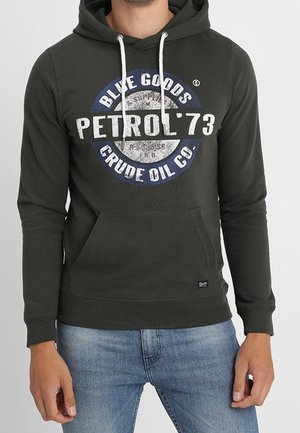 Mężczyzna w ciemnozielonej bluzie z kapturem, z białymi sznurkami i logo w stylu vintage „Petrol ’73 Blue Goods Crude Oil Co.”, w połączeniu z niebieskimi dżinsami.