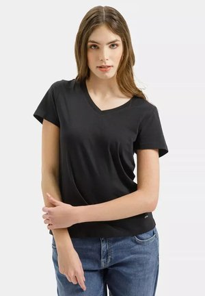 Młoda kobieta ubrana w czarną koszulkę z dekoltem w serek i niebieskie dżinsy, stojąca z założonymi na piersi ramionami na tle jasnego, jednolitego tła.