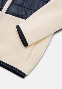 Light cream fleece jacket with dark navy padded chest panel. Features contrasting navy cuffs with stripes and an integrated zipper.