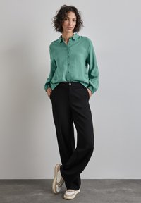 Chemise boutonnée vert menthe à manches longues, assortie à un pantalon large noir et des baskets beiges avec des accents blancs.