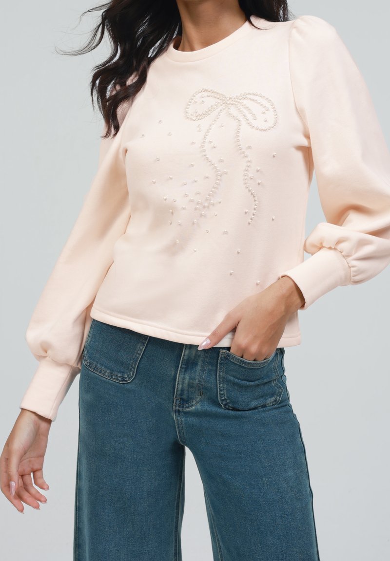 Femme portant un haut à manches longues rose clair avec un motif de nœud en perles et un jean bleu, une main dans la poche, debout devant un fond uni.