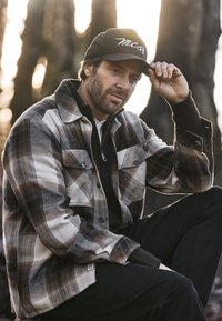 Camisa de franela a cuadros en beige y marrón, con botones y cuello. Debajo, un suéter oscuro con cremallera. Persona con gorra negra.