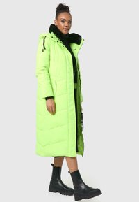 Neon groene pufferjas met zwart nepbont kraag, lange ontwerp, gestikte textuur en zijzakken, gecombineerd met zwarte enkelboots.