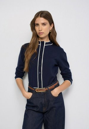 Mujer con el cabello largo y castaño que lleva un cárdigan azul marino abotonado con ribete blanco, vaqueros oscuros y un cinturón marrón, de pie con las manos en los bolsillos.