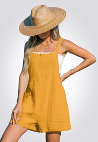 Vrouw in een breedgerande strohoed, mosterdgele mouwloze romper met voorzakken, witte crop top en gouden sieraden, poserende met haar hand op haar heup.