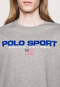 Šedé bavlněné tričko s výrazným modrým nápisem „POLO SPORT” a „RALPH LAUREN“ a malou grafikou americké vlajky pod ním.
