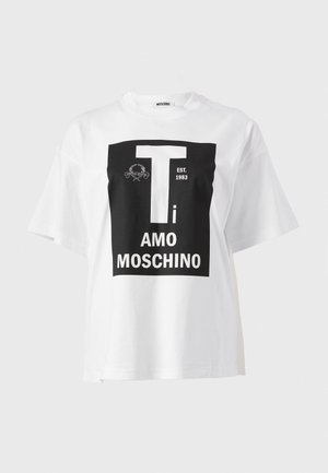Une femme porte un T-shirt blanc à motif graphique, un jean à jambes larges, des sandales noires et un sac à main noir sur un fond de studio uni.