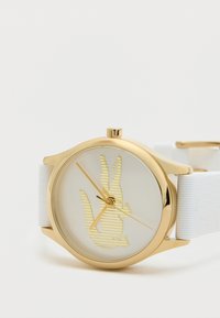 Zelta tonis pulkstenis ar baltu silikona jostu. Apaļa korpuss, skaidra ciparnīca ar dzeltenu svītru dizainu un zeltainām rokām.